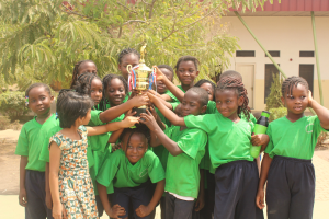 Gruppe von Kindern, die eine Trophäe in einer Schulumgebung mit Bäumen und einem Gebäude im Hintergrund halten.