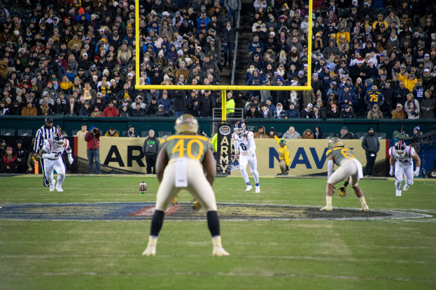 Fußballspieler in Sportkleidung und Helmen konkurrieren in einem Stadion, während Zuschauer von den Tribünen zuschauen, mit sichtbaren Stadionanzeigen und strukturellen Elementen.