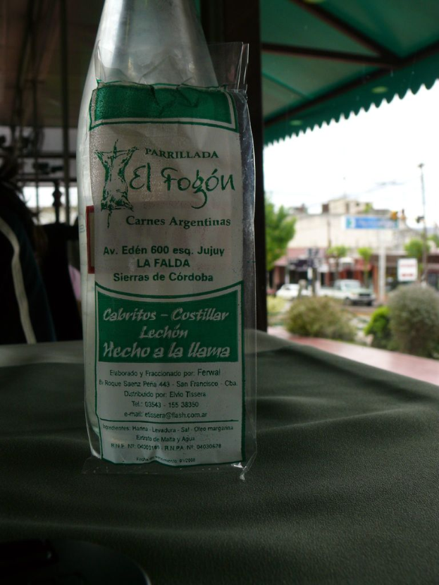 Eine Flasche mit beschrifteter Text auf einem Tisch, mit einer Säule und Dach dahinter, sowie einem Gebäude, einem Fahrzeug, Hecken, einem Pfahl und Himmel im Hintergrund.