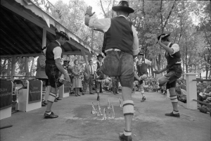 Eine Gruppe von Männern in traditioneller bayerischer Tracht tanzt auf einer Bühne vor einem gemischten Publikum aus sitzenden und stehenden Menschen, mit Bäumen und einer Hütte im Hintergrund, dargestellt in Schwarz-Weiß.