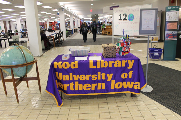 Eine Ausstellung mit einem Tisch, auf dem verschiedene Landesflaggen, eine Box, Papiere und ein Banner liegen, eine Kugel links daneben, ein Anschlagbrett rechts mit ein paar Leuten daneben und mehreren Tischen und Stühlen im Hintergrund.