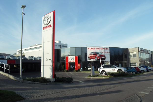 Ein Toyota-Händler mit parkenden Autos davor, umgeben von Gebäuden, Werbetafeln, Straßenlaternen, Straßenfahrzeugen auf der Straße, Büschen, Bäumen und einem bewölkten Himmel im Hintergrund.