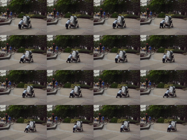 Ein Kind schiebt einen Wagen auf der Straße, mit zwei Personen, die links Fahrräder halten, Bäumen im Hintergrund und einem Gebäude links