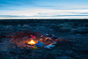 Ein Lagerfeuer mit Menschen, die darauf sitzen und liegen, mit Mützen tragend, mit Taschen und anderen Gegenständen auf dem Boden, umgeben von Bäumen und Himmel.