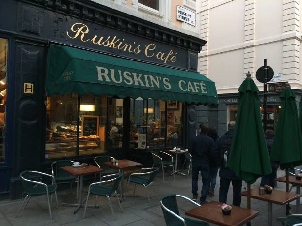 Eine Gruppe von Menschen steht vor einem Cafe mit einer Tafel, auf der 'Ruskins Cafe' steht.