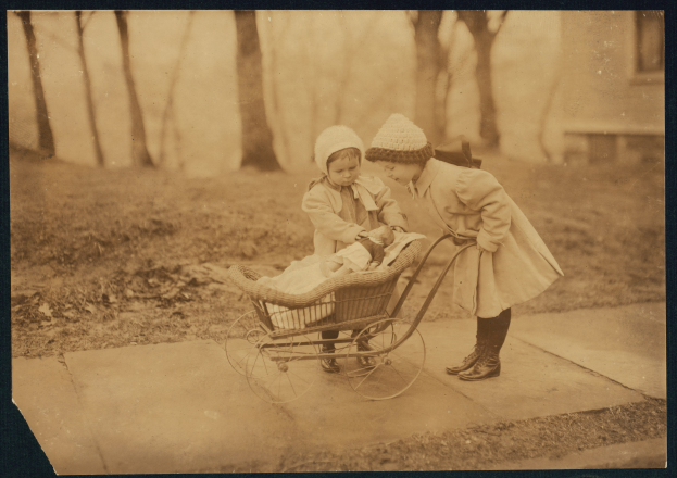 Zwei Kinder in Mänteln und Mützen schieben einen Kinderwagen, mit Bäumen, einem Gebäude und Gras im Hintergrund.