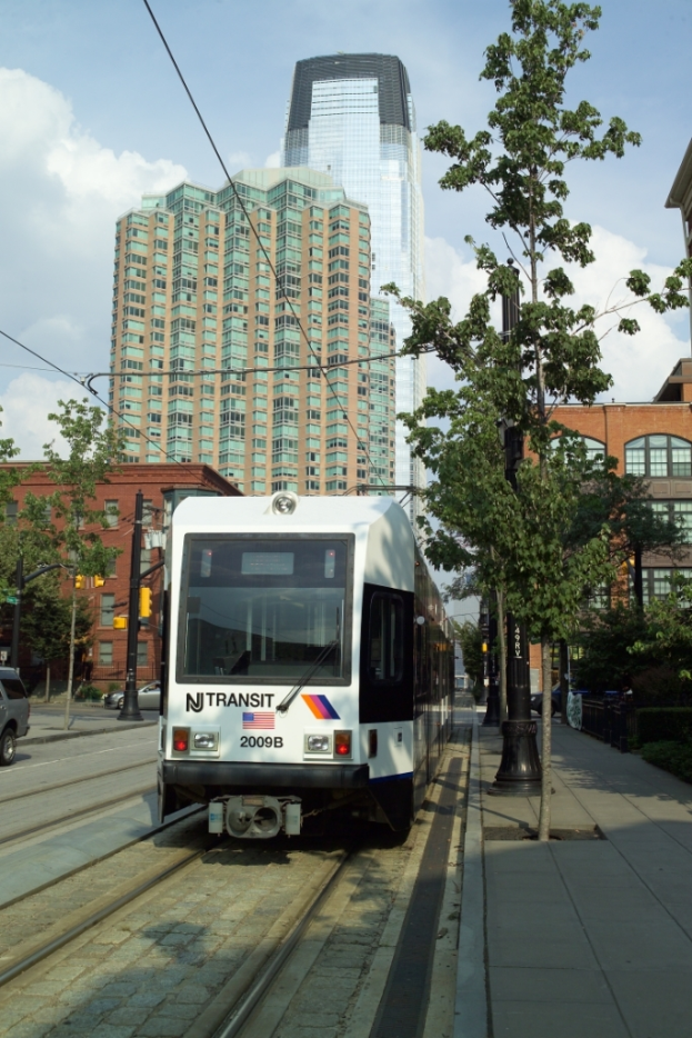 Ein Zug auf Schienen mit elektrischen Kabeln darauf, umgeben von Bäumen, Pfählen und Gebäuden, mit einem Wolkenkratzer und Himmel im Hintergrund.