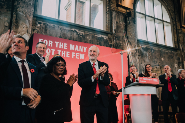 Eine Gruppe von Menschen klatscht feiernd vor einem Podium mit einer Tafel, die Text anzeigt, einer Wand mit einem Banner dahinter und verstreuten Stühlen im Raum.