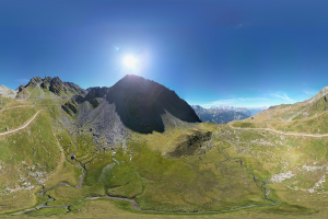 Panoramischer Blick auf ein Gebirgszug mit grünem Gras, einer gewundenen Straße und einem hellblauen Himmel unter einer strahlenden Sonne.