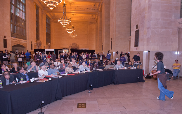 Ein Konzertbild mit einer Menge Menschen, einem Mann, der rechts Gitarre spielt und singt, einer Wand und Fenstern im Hintergrund, Lichtern oben und einem Boden unten.