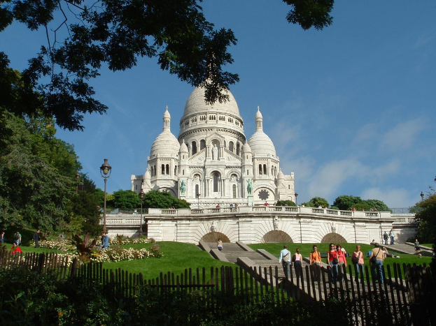 Ein weißes Gebäude umgeben von einem Zaun, Gras, Bäumen und Menschen mit Stangen.