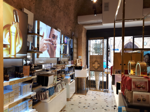 Innenansicht eines Kosmetikgeschäfts mit verschiedenen Produkten auf Gestellen, Bildschirmen an den Wänden, Deckenleuchten, einer Klimaanlage und Glastüren, die eine Person, ein Auto und ein Gebäude draußen zeigen.