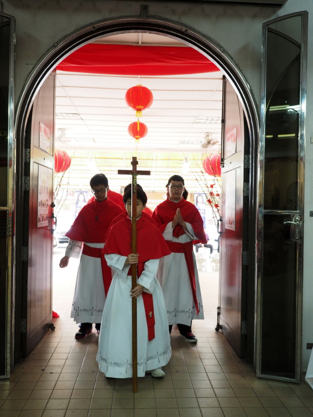 Menschen mit einem Kreuz in einer Kathedrale spazieren, mit Türen links und rechts und Blumen und dekorativen Elementen im Hintergrund.