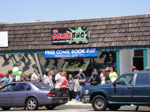 Eine Gruppe von Menschen vor einem Gebäude mit Glastüren und einem Dach, zwei Autos auf der Straße, grüne Luftballons, eine Fahne und ein Namensschild auf dem Dach unter einem klaren Himmel.