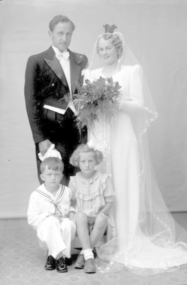 Schwarzes und weißes Foto einer Braut in einem weißen Kleid mit einem Strauß, Bräutigam, der daneben lächelt, und zwei Kindern, die vor ihnen auf dem Boden sitzen.