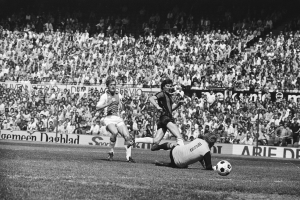 Schwarze und weiße Fotografie von Männern, die auf einem Feld Fußball spielen, mit Zuschauern im Hintergrund und einem Banner mit der Aufschrift "Bundesliga 1978-79 Ajax 1-1 Hannover 96".