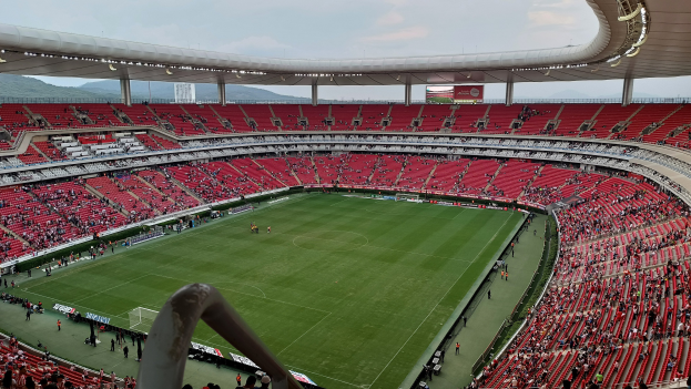 Ein großes Stadion voller Zuschauer bei einem Fußballspiel, mit Hügeln und einem klaren blauen Himmel im Hintergrund.