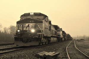 Ein Zug auf Schienen mit Autos, Strommasten, Bäumen und Himmel im Hintergrund.