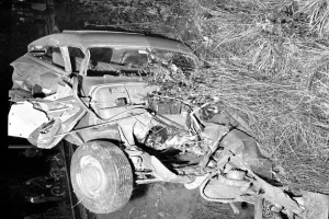 Ein Schwarz-Weiß-Foto eines schwerbeschädigten Autos am Straßenrand mit einer zerbeulten Front, zersplitterten Fenstern, einem Baum im Hintergrund und verstreuten Gegenständen rund um die Unfallstelle.