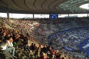Große Zuschauermenge in einem Stadion bei einem Fußballspiel, mit einer Bühne auf der rechten Seite, Fahnen, Stangen, einem Bildschirm und dem Allianz Arena in München, Deutschland im Hintergrund.