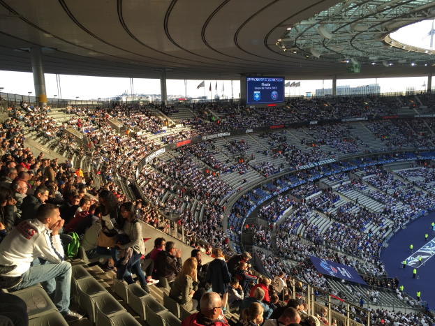 Große Zuschauermenge in einem Stadion bei einem Fußballspiel, mit einer Bühne auf der rechten Seite, Fahnen, Stangen, einem Bildschirm und dem Allianz Arena in München, Deutschland im Hintergrund.