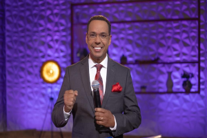 Stephen Colbert steht auf einer Bühne und hält ein Mikrofon in der Hand, trägt einen Anzug, eine Krawatte und eine Brille, während im Hintergrund ein Scheinwerfer und ein Ständer zu sehen sind.