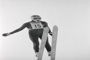 Ein Skispringer in der Luft, der einen Helm, eine Brille, Handschuhe und Schuhe trägt und Ski-Bretter hält.