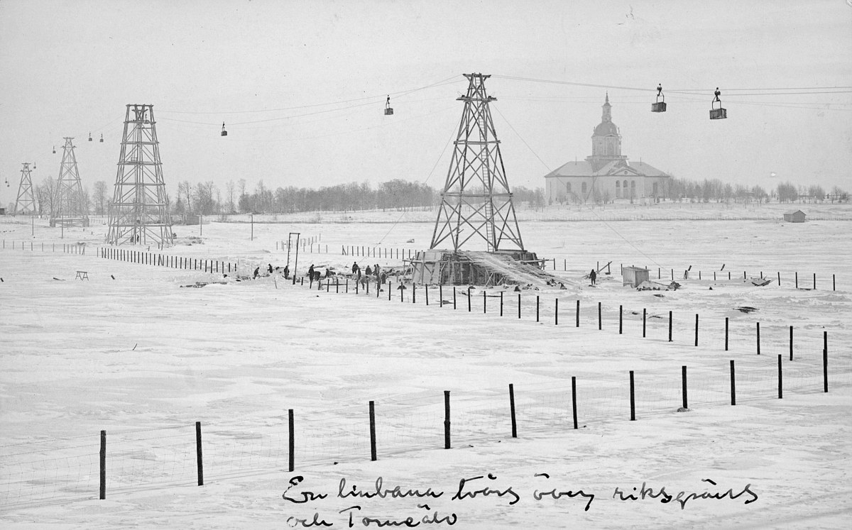 Schwarze-Weiß-Foto eines Skilifts in einer verschneiten Wiese mit Stützpfosten, Oberleitungen, Bäumen und einem Gebäude im Hintergrund, mit Text unten.