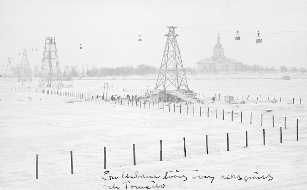 Schwarze-Weiß-Foto eines Skilifts in einer verschneiten Wiese mit Stützpfosten, Oberleitungen, Bäumen und einem Gebäude im Hintergrund, mit Text unten.