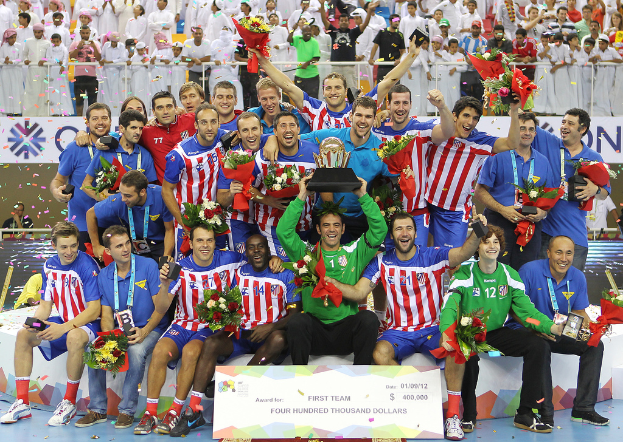 Eine Gruppe von Menschen in Sportkleidung, einige halten Auszeichnungen, Blumen und andere Gegenstände, steht vor einer Menge mit Geländern und Schildern im Hintergrund.