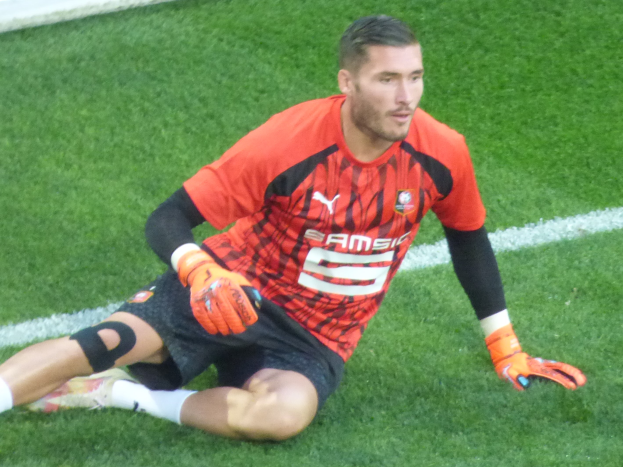 Ein Füßballspieler in rotem T-Shirt, schwarzen Shorts, weißen Socken und orangefarbenen Baseballhandschuhen sitzt auf dem Boden mit händen auf den Knien.