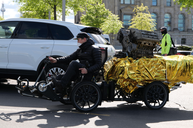 Ein Mann im Rollstuhl mit einem großen Motor auf dem Rücken, umgeben von Fahrzeugen auf einer Straße mit Bäumen, Gebäuden, Polen und einem klaren blauen Himmel im Hintergrund, trägt eine schwarze Jacke, eine Mütze und hält ein Objekt in der Hand.