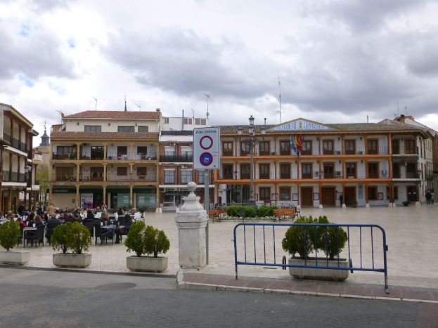 Ein belebter Stadtplatz mit Menschen auf Stühlen und Stehenden, umgeben von Topfpflanzen, Metallabsperrungen, Straßenmasten mit Flaggen, einem Schild, Gebäuden und einem bewölkten Himmel.