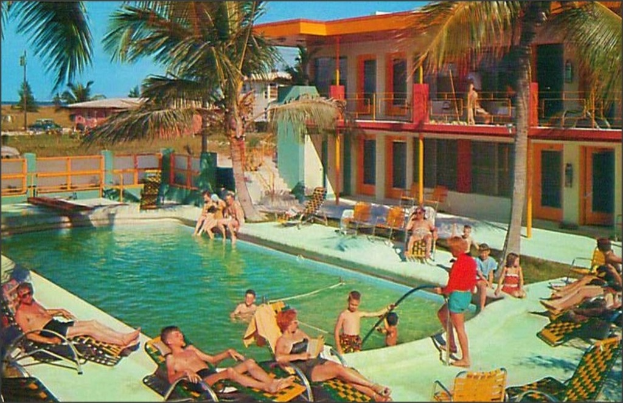 Ein Schwimmbad mit Menschen auf Stühlen im Vordergrund, Gebäuden und Bäumen im Hintergrund und sichtbaren Autos.