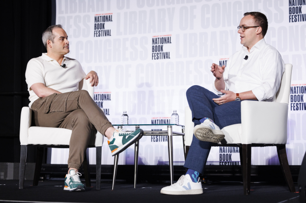 Zwei Männer sitzen auf Stühlen auf einer Bühne mit einem Tisch dazwischen, der zwei Wasserflaschen und ein Banner im Hintergrund zeigt, auf dem "National Book Festival" steht.