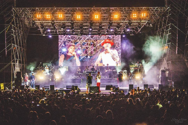 Eine Gruppe von Menschen auf einer hell erleuchteten Bühne in der O2 Arena in London, einige spielen Instrumente und andere halten Handys hoch, vor einem dunklen Hintergrund.