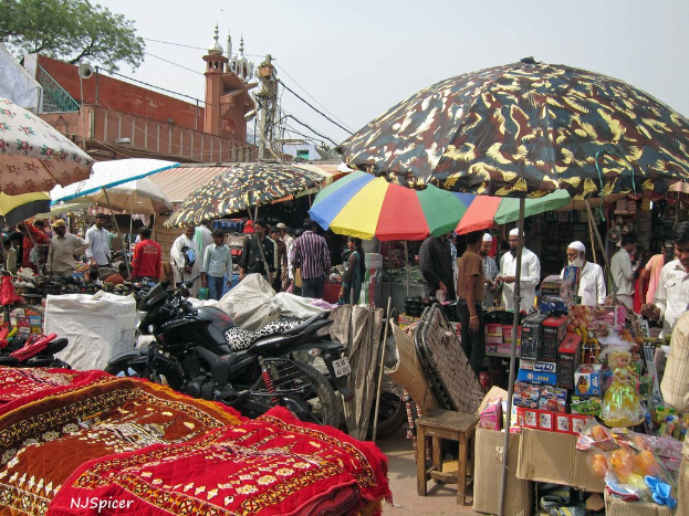 Eine belebte Szene mit Schirmen, Fahrzeugen, Matten, Kisten, einem Hocker, Menschen und verschiedenen anderen Gegenständen, mit einer Moschee, Mikrofonen, einem Strommast und Kabeln im Hintergrund.