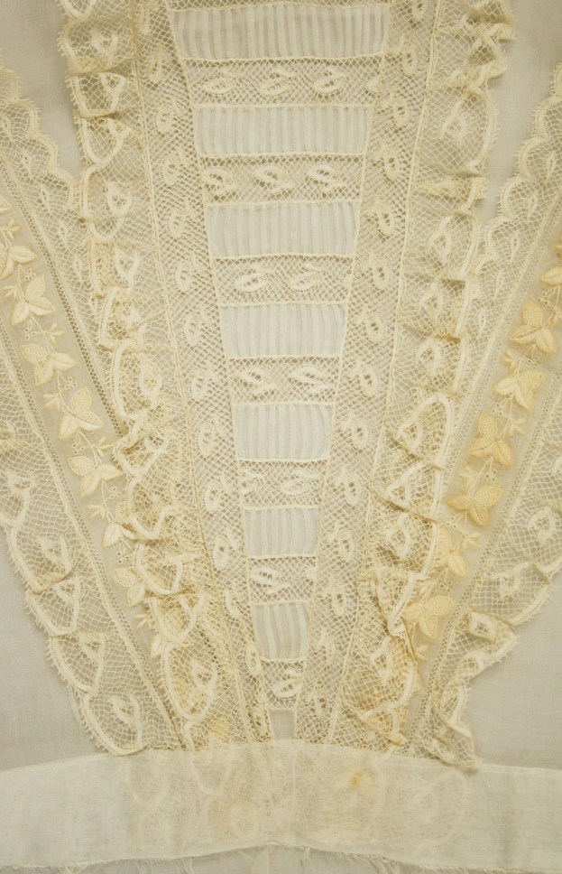 Nahaufnahme eines weißen Kleides mit zarter, detallierter Spitzenverzierung mit verschiedenen Formen und Mustern auf leichtem, luftigem Stoff.