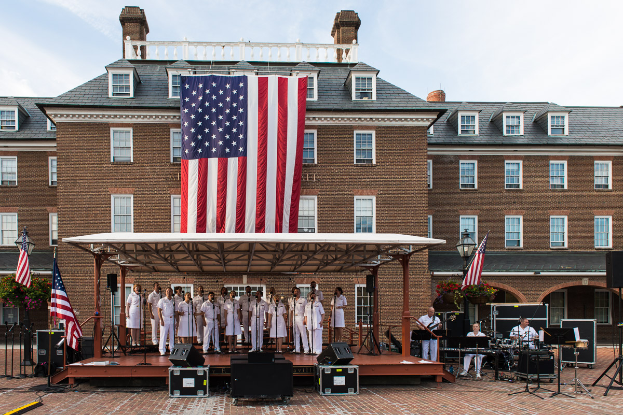 Eine Gruppe von Menschen auf einer Bühne vor einem Gebäude mit Fenstern, umgeben von Fahnen, Lautsprechern und Blumenpflanzen, die während eines Gedenkfeiertags-Konzerts auf dem Mount Vernon spielen.