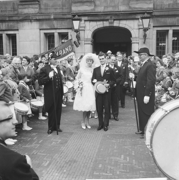 Ein Brautpaar geht eine Straße entlang vor einer Menge, einige halten Musikinstrumente, mit einem Gebäude und einem Banner im Hintergrund, alles in Schwarz-Weiß dargestellt.