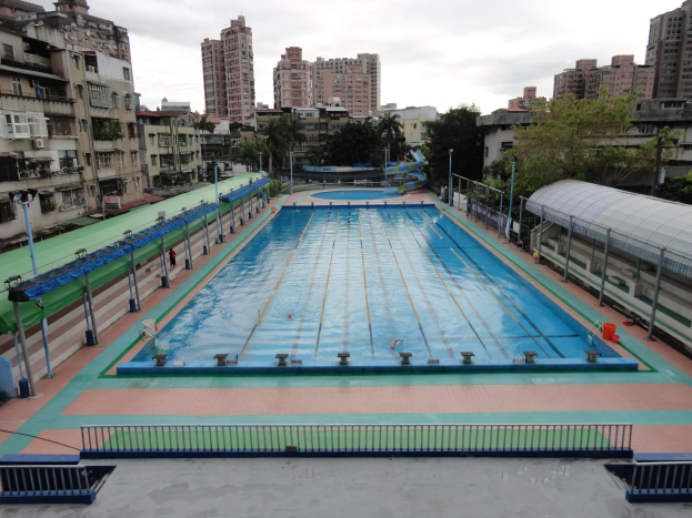 Großer städtischer Schwimmbadkomplex umgeben von Zäunen, Pfählen, Laternen, Bäumen und Gebäuden mit einem Schuppen auf der rechten Seite und Himmel im Hintergrund.