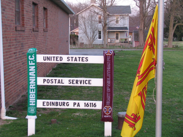 Ein grasbewachsener Bereich mit einem zentralen Mast, an dem Fahnen und ein "United States Postal Service"-Schild angebracht sind, neben einem Gebäude mit Fenstern unter einem klaren blauen Himmel mit Bäumen im Hintergrund.