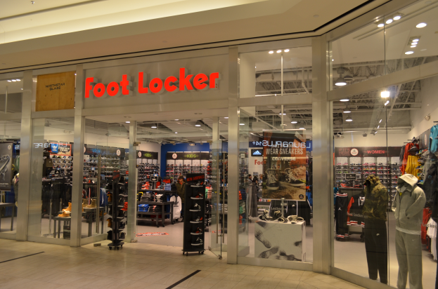 Ein Foot Locker Geschäft in einem Einkaufszentrum mit Glaswänden und -türen, Schaufensterpuppen in Kleidern, Regalen mit Schuhen und anderen Waren sowie Deckenbeleuchtung.