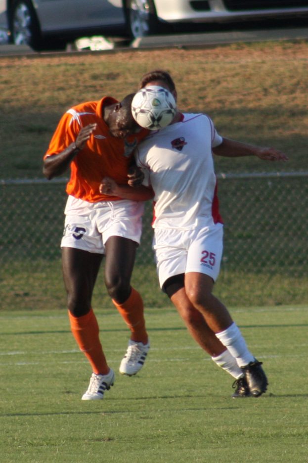 Zwei Spieler auf einem umzäunten Platz, einer im orangen T-Shirt schlägt den Ball mit dem Kopf, mit Autos und Gras im Hintergrund.