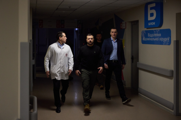 Eine Gruppe von Männern geht einen hell erleuchteten Krankenhausflur entlang, mit einer Tür auf der rechten Seite und Informationsschildern auf der linken Seite, die besorgt aussehen.