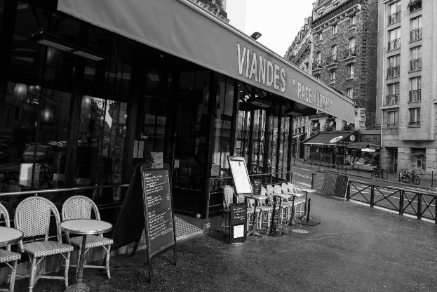 Ein Schwarz-Weiß-Bild einer StraßenSzene mit einem Geschäft und Stühlen in der Mitte, einem Gebäude auf der rechten Seite, einer Straße und Fahrrädern.