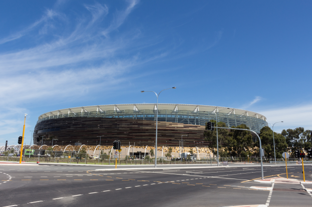 Ein großes Stadion mit starkem Verkehr davor, umgeben von Straßenmasten, Laternen, Ampeln, Schildern, Geländern, Bäumen und einem bewölkten Himmel.