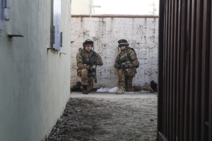 Eine Szene mit einer Wand und einem Fenster auf der linken Seite, einer eisernen Wand auf der rechten Seite, zwei Soldaten und Erde in der Mitte und Gebäuden, einem Strommast und einer weiteren Wand im Hintergrund.
