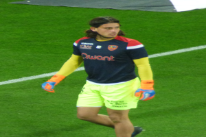 Ein Mann in einem blauen Hemd und gelben Shorts läuft auf einem Fußballfeld und hat die Arme ausgestreckt und einen entschlossenen Gesichtsausdruck, mit einem Tuch oben auf dem Bild.