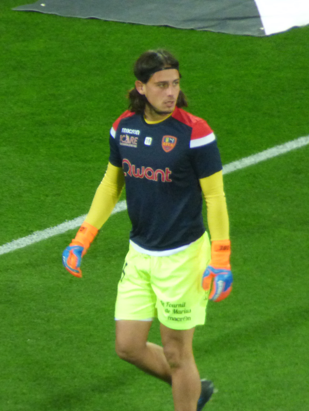 Ein Mann in einem blauen Hemd und gelben Shorts läuft auf einem Fußballfeld und hat die Arme ausgestreckt und einen entschlossenen Gesichtsausdruck, mit einem Tuch oben auf dem Bild.